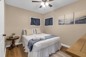 Quarto com cama, mesa de cabeceira, ventoinha no teto e janela com cortinas.