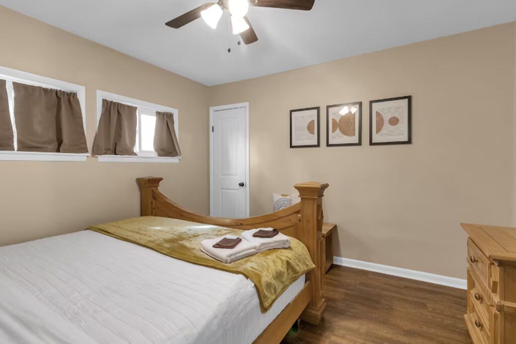 A bedroom with a bed, a wooden headboard, a ceiling fan, a window with curtains, and a door.