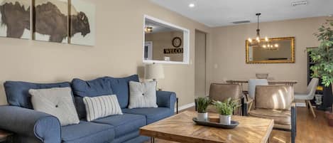 A living room with a blue sofa, a wooden coffee table, and a dining area with chairs.