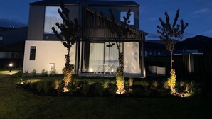 Une maison moderne à deux étages, dotée d’une grande fenêtre, d’une petite porte et d’un patio aménagé avec du mobilier d’extérieur.