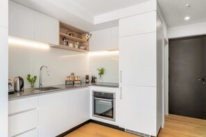 Una cocina compacta con armarios blancos, horno integrado, lavamanos y encimera con tetera y cafetera.