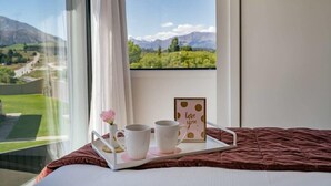 Une chambre avec une grande fenêtre, un lit recouvert d’une couverture marron et un plateau sur lequel se trouvent deux tasses blanches et une petite fleur rose.