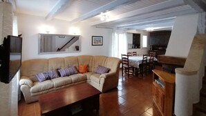Uma sala de estar com um sofá seccional bege, uma mesa de centro de madeira e uma área de refeições com toalha xadrez e cadeiras de madeira.