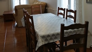 Uma área de jantar com mesa posta para quatro pessoas, cadeiras de madeira, um sofá e uma televisão na parede.