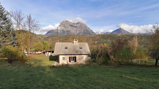 Uma pequena casa com telhado inclinado, cercada por um campo gramado e árvores.