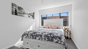 A bedroom with a bed, a window with bars, a painting on the wall, and a staircase leading to another room.