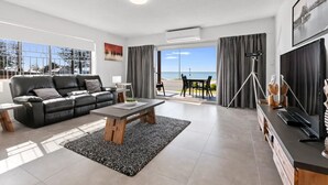 A modern living room with a black leather sofa, a wooden coffee table, and a dining area with a table and chairs. There is a large window with a view of the sea and a balcony with a table and chairs.