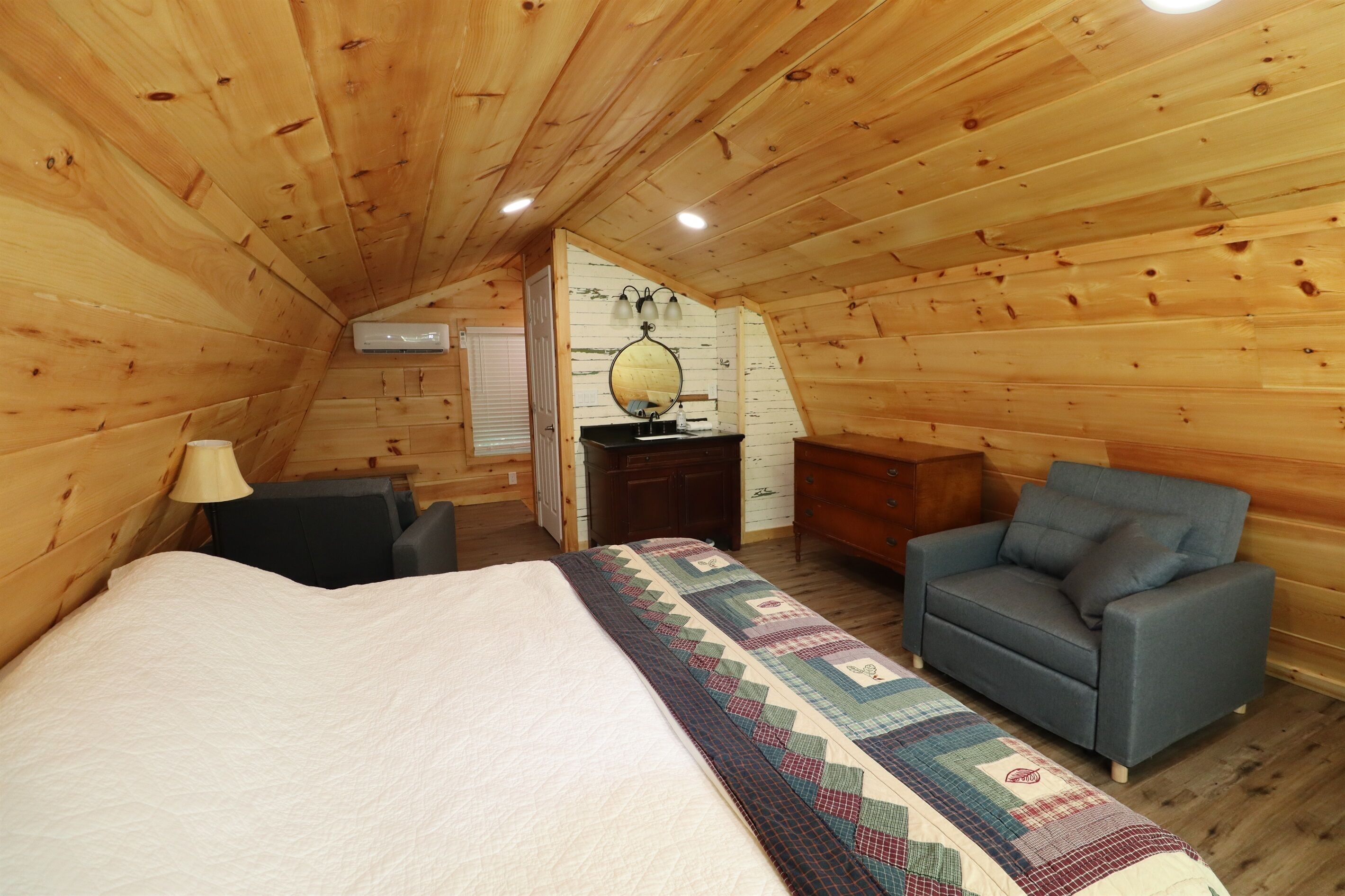 A cozy attic bedroom with a bed, a blue armchair, a dresser, and a mirror on the wall.
