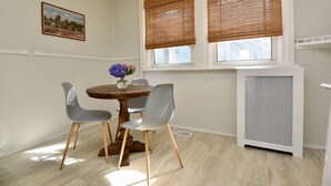 A dining area with a round table, two chairs, a vase with flowers, and a window with blinds.