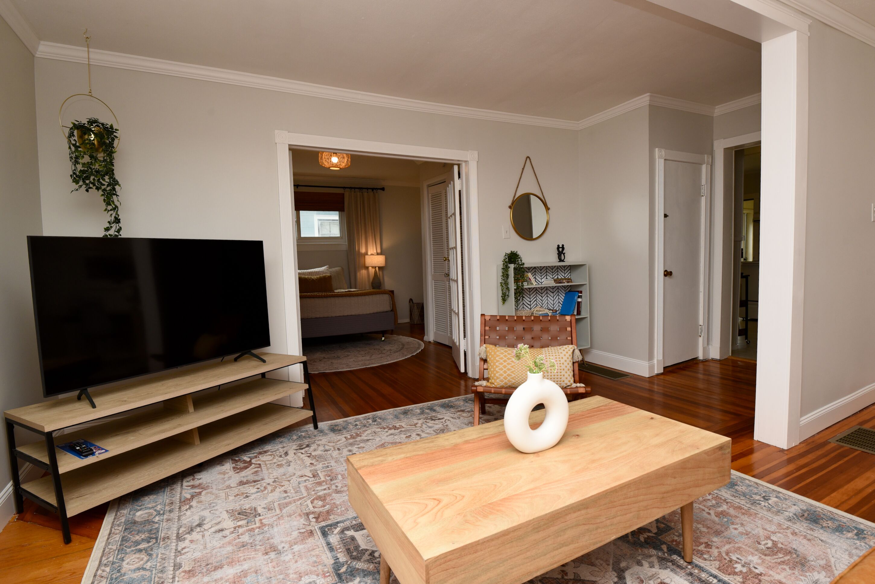 Una sala de estar con una televisión de pantalla plana sobre un soporte de madera, una mesa de centro con un florero, un tapete estampado y un sillón con cojín.
