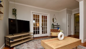 A living room with a wooden coffee table, a TV on a wooden stand, a patterned rug, a door with glass panels, and a small plant on the wall.