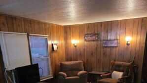 Une pièce chaleureuse avec des murs en bois, un fauteuil marron, un fauteuil à carreaux, un canapé et un téléviseur posé sur un meuble en bois.