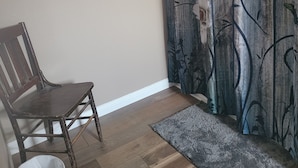 Une pièce avec une chaise en bois, une poubelle blanche, un tapis gris et un rideau à motifs floraux.