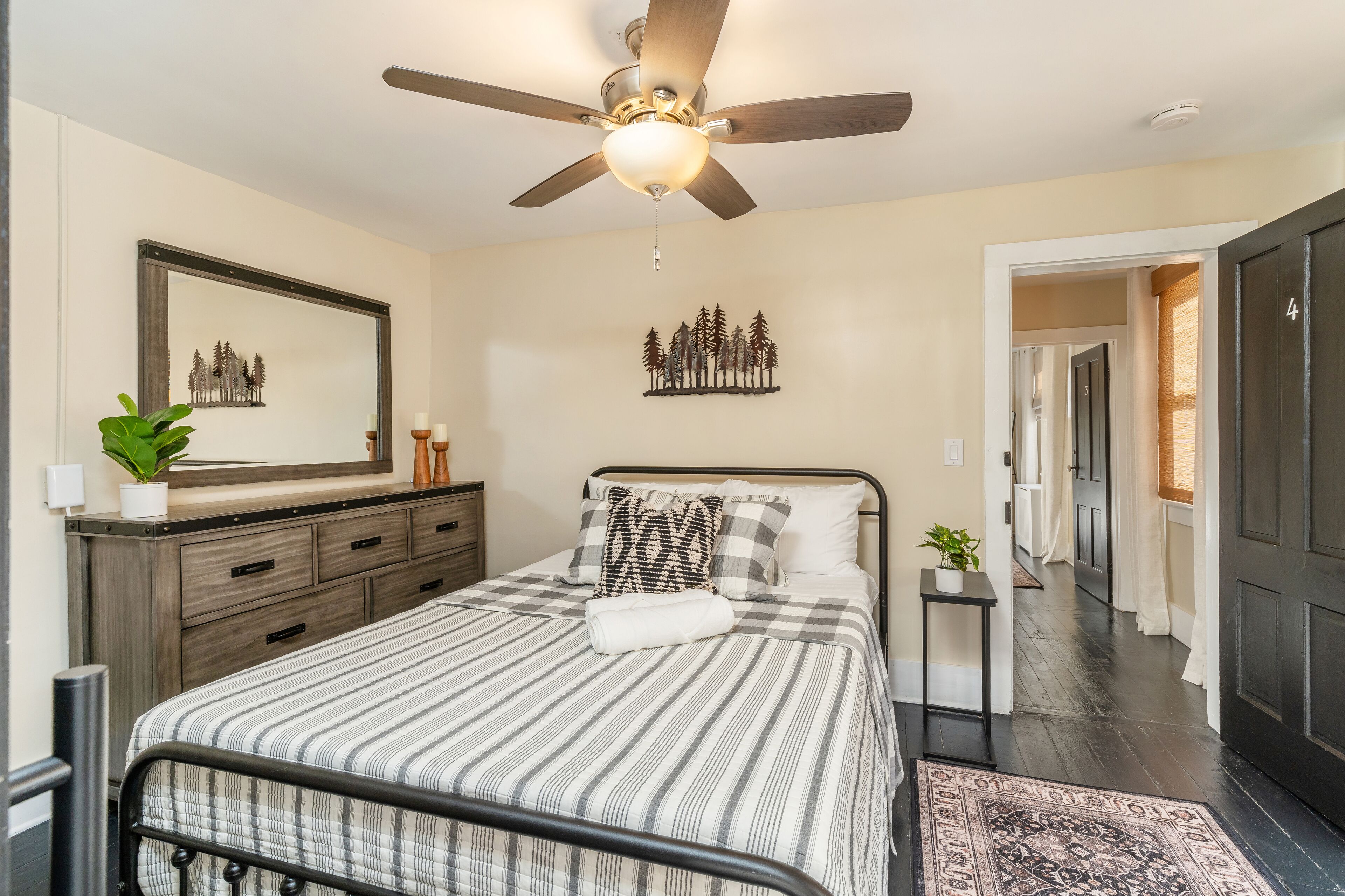 A bedroom with a bed, a ceiling fan, a nightstand, a dresser, a mirror, and a hallway leading to another room.