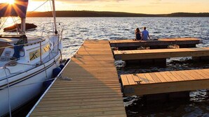 Un velero amarrado a un muelle de madera, con dos personas sentadas en él, contemplando un cuerpo de agua tranquilo durante la puesta de sol.