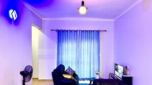 A modern living room with a blue sofa, a black armchair, a standing fan, a TV, and a blue curtain. - C&A Orchid Apartments (Kampala)