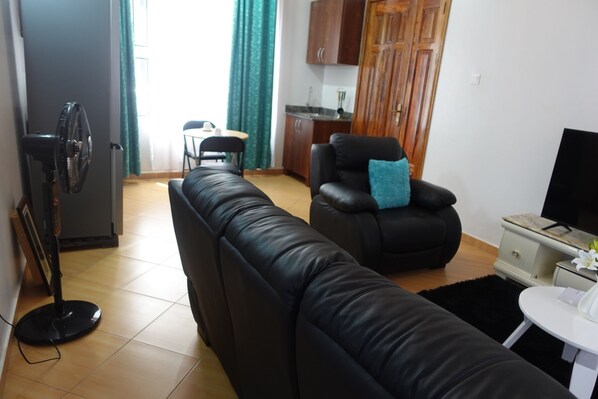 A living room with a black leather sofa, a black armchair with a blue pillow, a standing fan, a TV on a white table, and a kitchen area in the background. - C&A Orchid Apartments (Kampala)