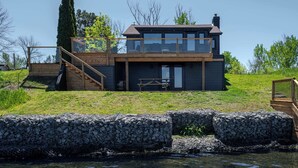 Una casa moderna de dos pisos con un balcón de madera, una escalera y vistas a un cuerpo de agua.