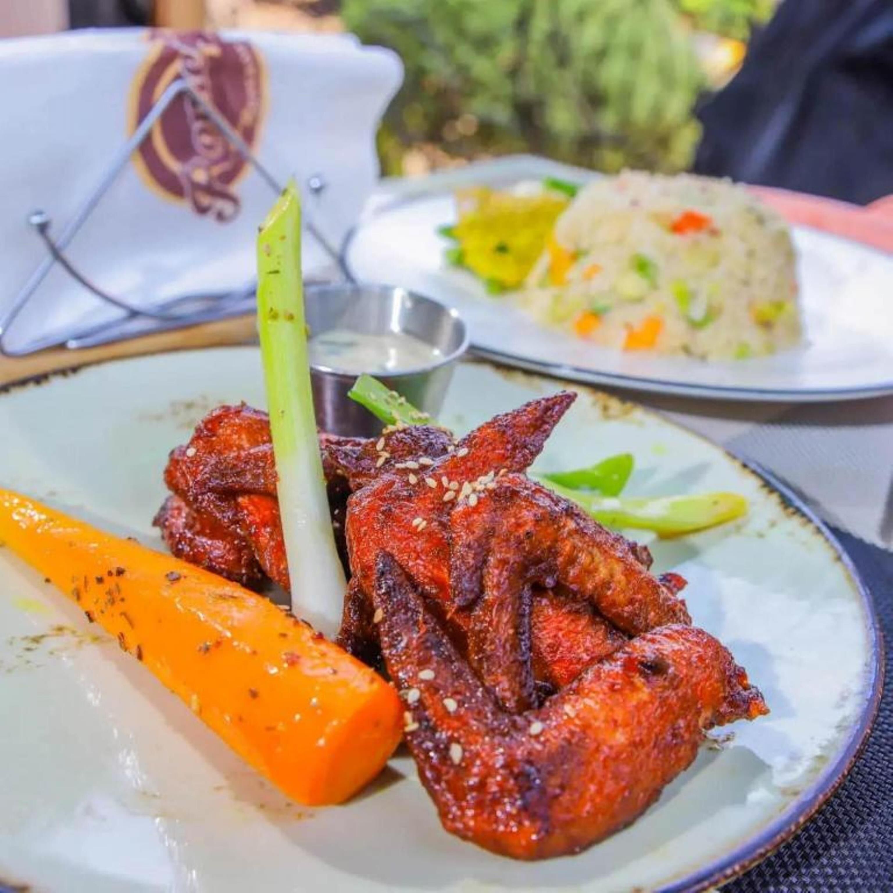 A plate of grilled chicken wings with a side of carrot and a small bowl of sauce.