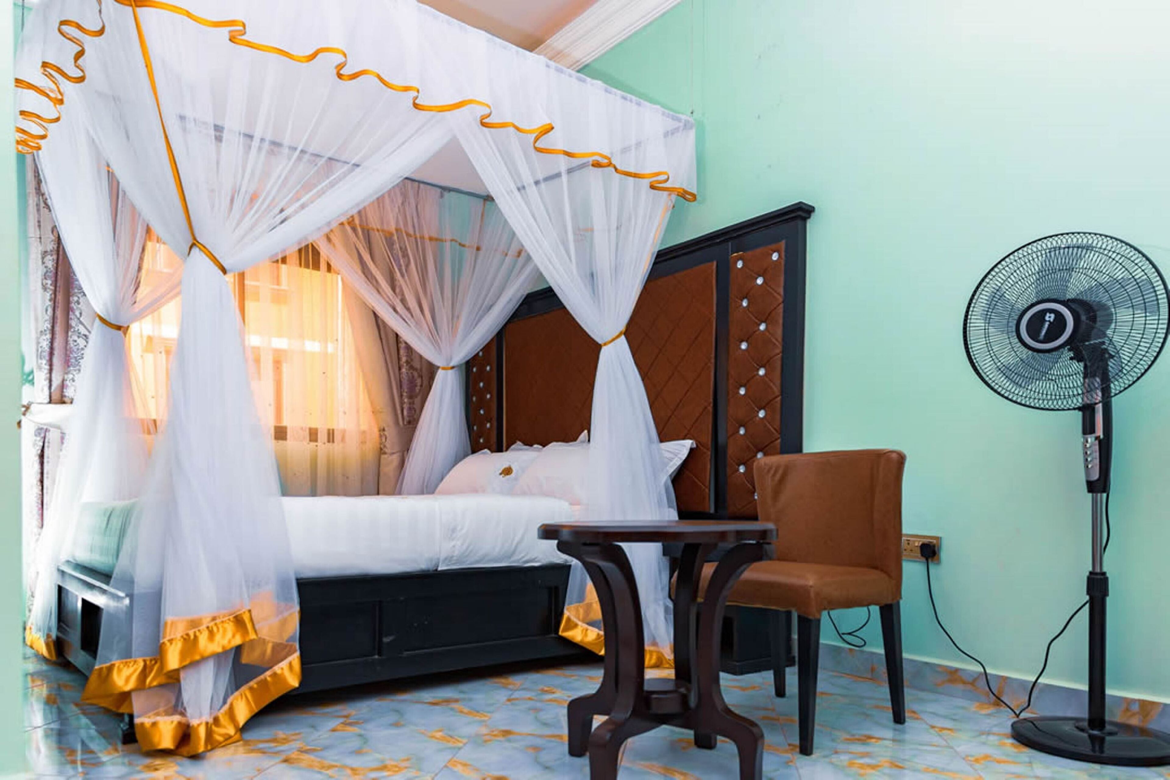 A bedroom with a bed under a white canopy, a bedside table, a chair, and a standing fan.