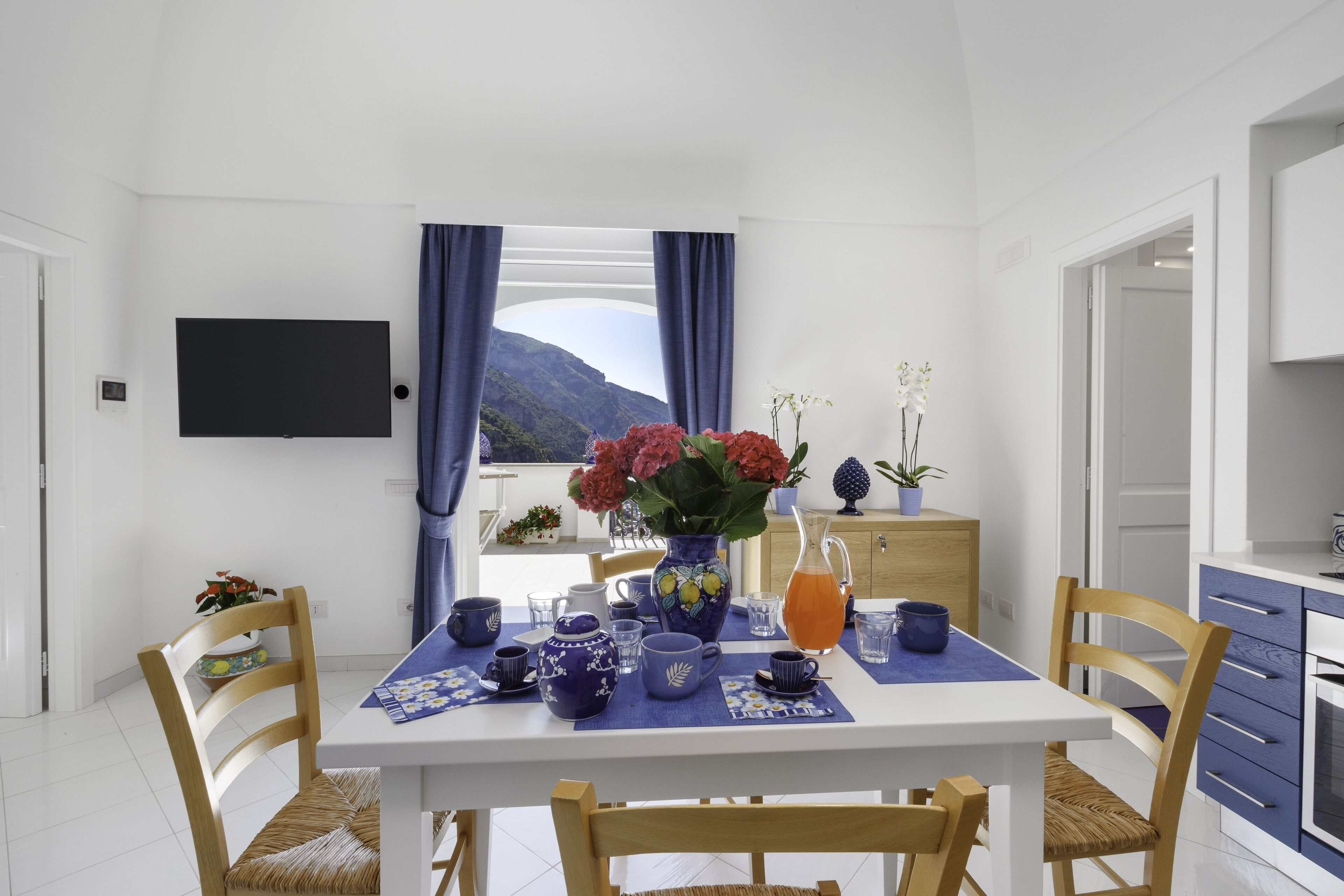 A dining area with a table set for four, a vase with flowers, and a view of mountains through the window.