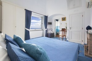 A bedroom with a large bed, a window with curtains, a small desk, and a bathroom visible through an archway. - Villa Settemari (Positano)