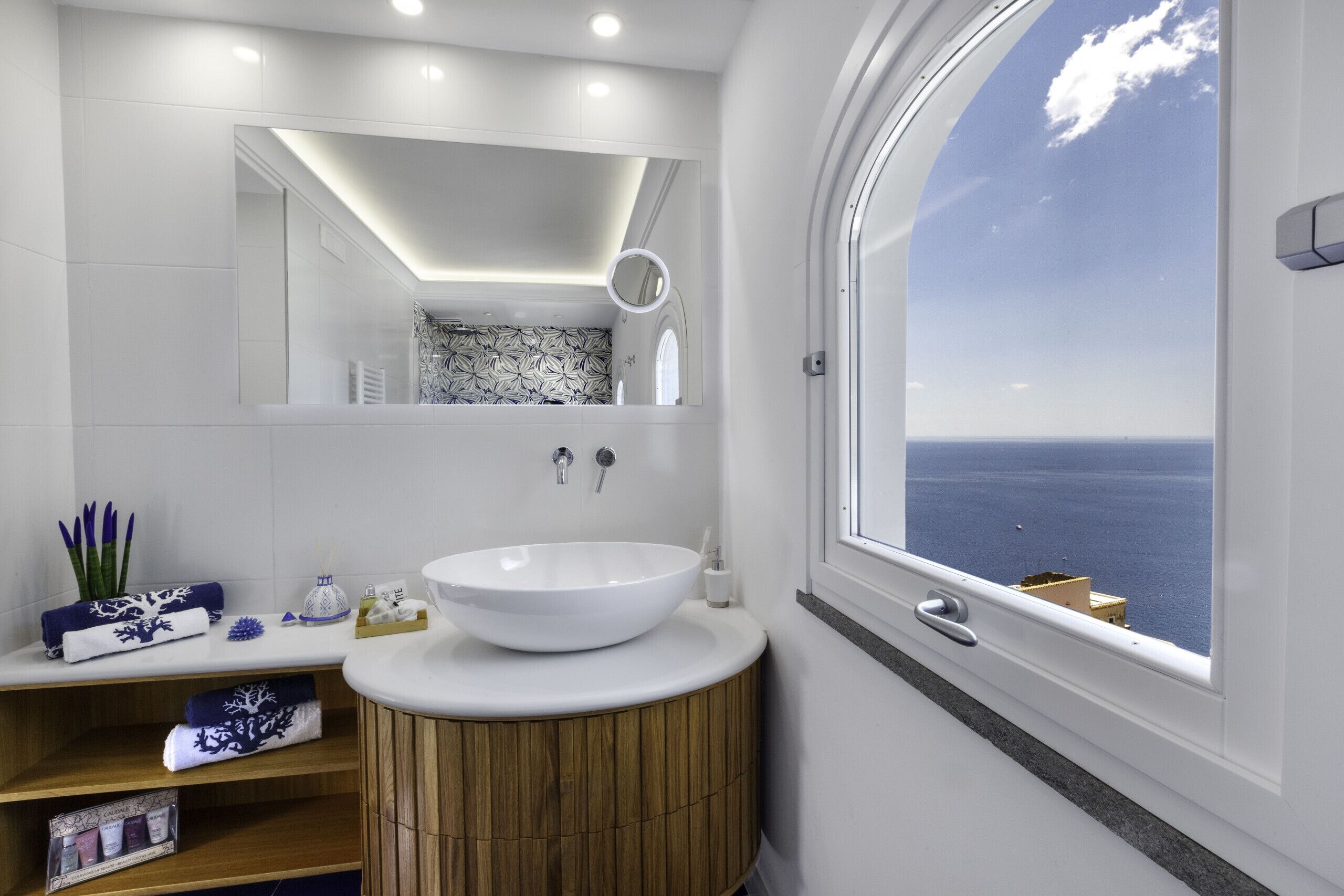 A modern bathroom with a large mirror, a round window with a view of the sea, a wooden vanity, and a white sink.