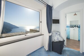 Een kamer met een groot raam met uitzicht op de kust, een klein bed met een kussen, een aan de muur bevestigde televisie, en een keukenhoek met blauwe en witte tegels.