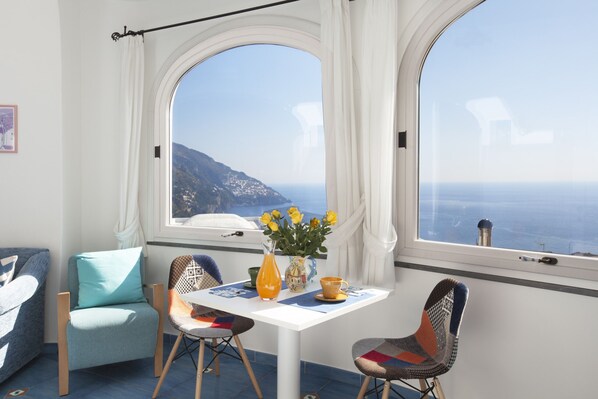 Een kamer met een tafel en stoelen, een raam met uitzicht op zee en een vaas met bloemen.