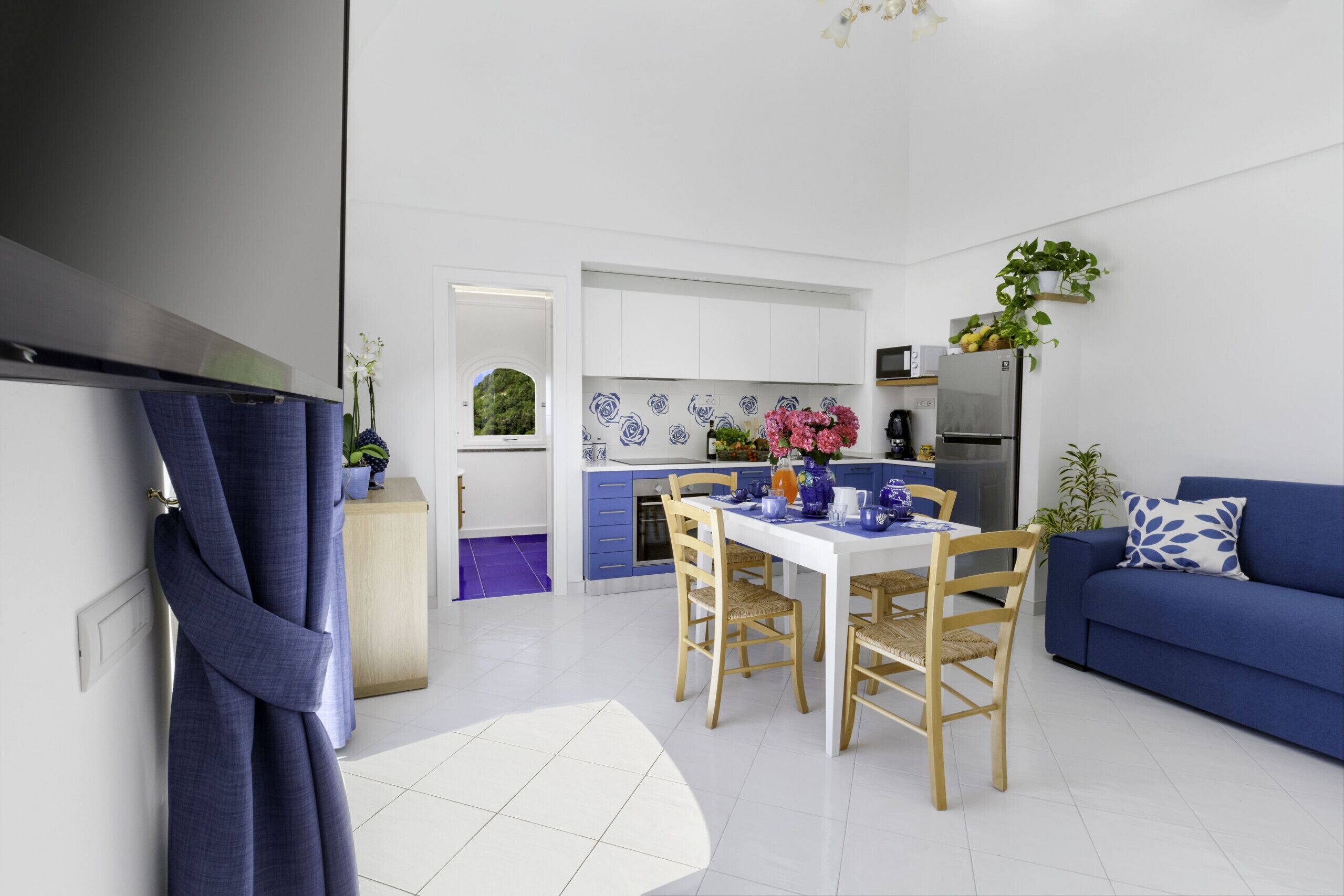 A modern kitchen with white cabinets, a blue countertop, and a refrigerator. There is a dining table with chairs and a blue sofa in the living area.