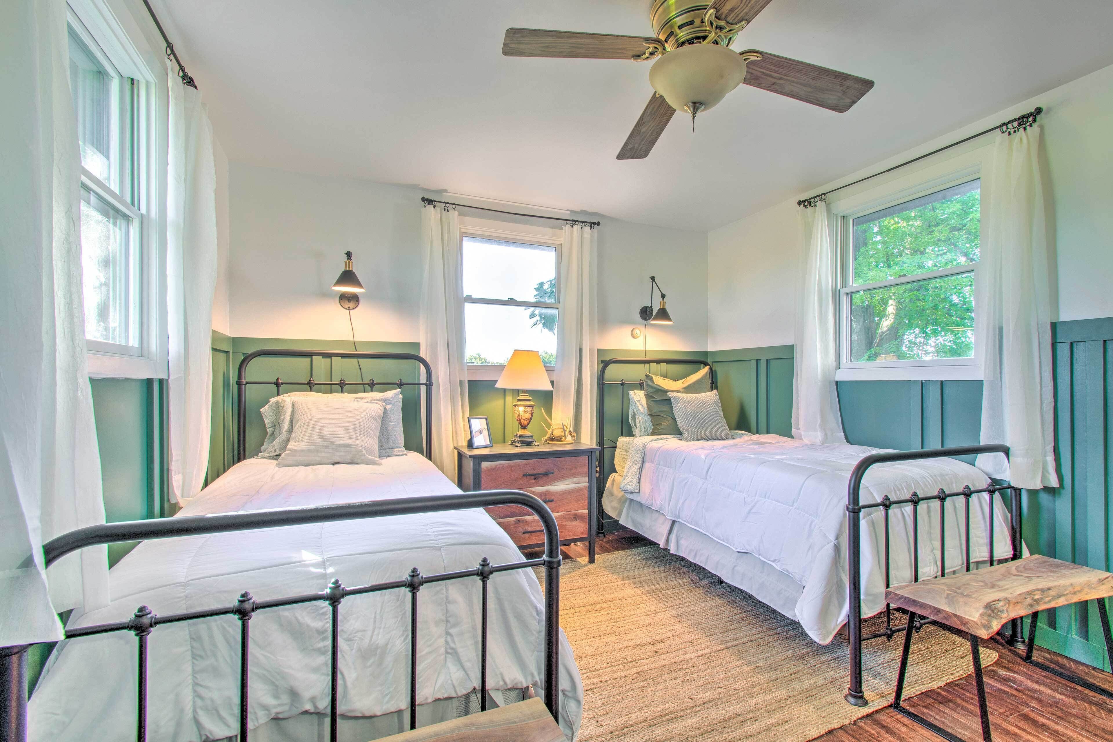 A room with two metal beds, a wooden bench, a ceiling fan, and a window with curtains.