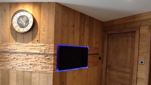 Une pièce aux murs recouverts de lambris en bois, avec une horloge murale, un téléviseur à écran plat doté de diodes électroluminescentes bleues et une porte en bois.
