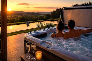 Uma banheira de hidromassagem com duas pessoas, um deck de madeira e vista para um lago e colinas ao pôr do sol.