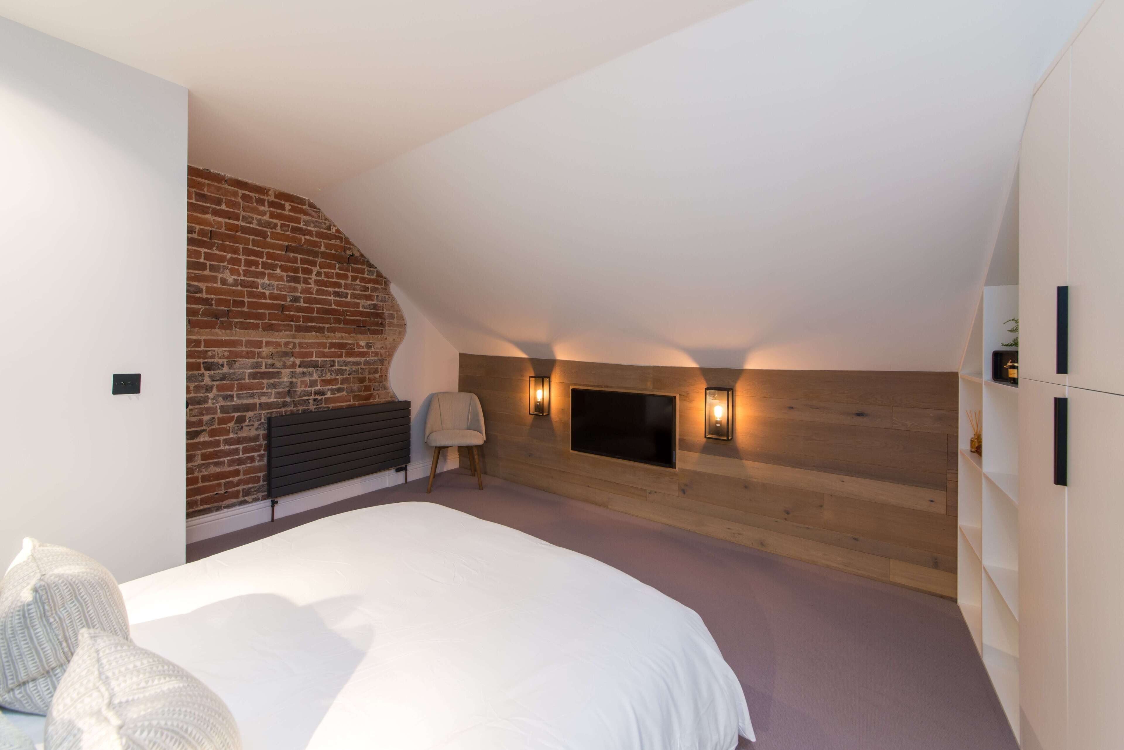 A bedroom with a sloped ceiling, a brick wall, a bed, a chair, a TV, and built-in shelves.