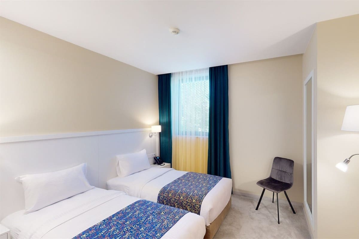 A hotel room with two beds, a chair, a desk, a lamp, and a window with curtains.