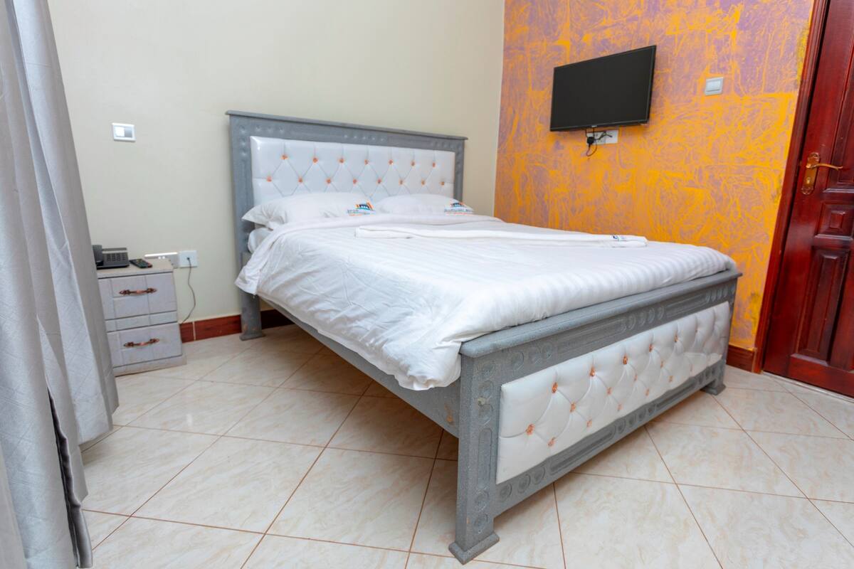 A neatly made bed with a grey headboard and white bedding, a bedside table with a lamp, and a TV mounted on the wall.