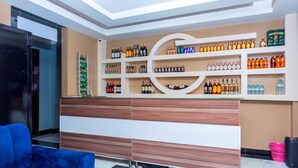 A reception area with a wooden and white reception desk, shelves with various liquor bottles, a blue sofa, and a pink basket. - Spotlight Motel (Kampala)