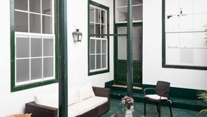 A room with a sofa, a chair, a table with a vase, and large windows with green frames.