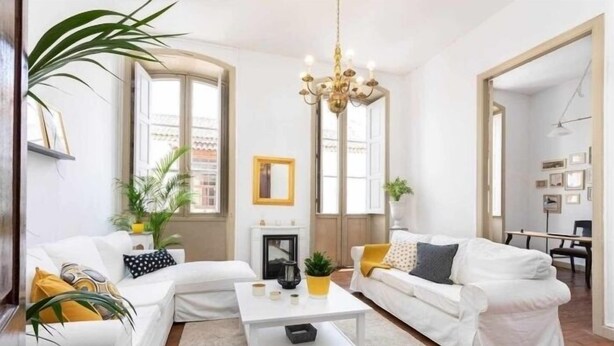 A modern living room with white sofas, a white coffee table, and a chandelier. There are potted plants and a fireplace.