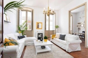 Ein modernes Wohnzimmer mit weißen Sofas, einem weißen Couchtisch und einer Kronleuchter. Es gibt Blumentöpfe und einen Kamin.