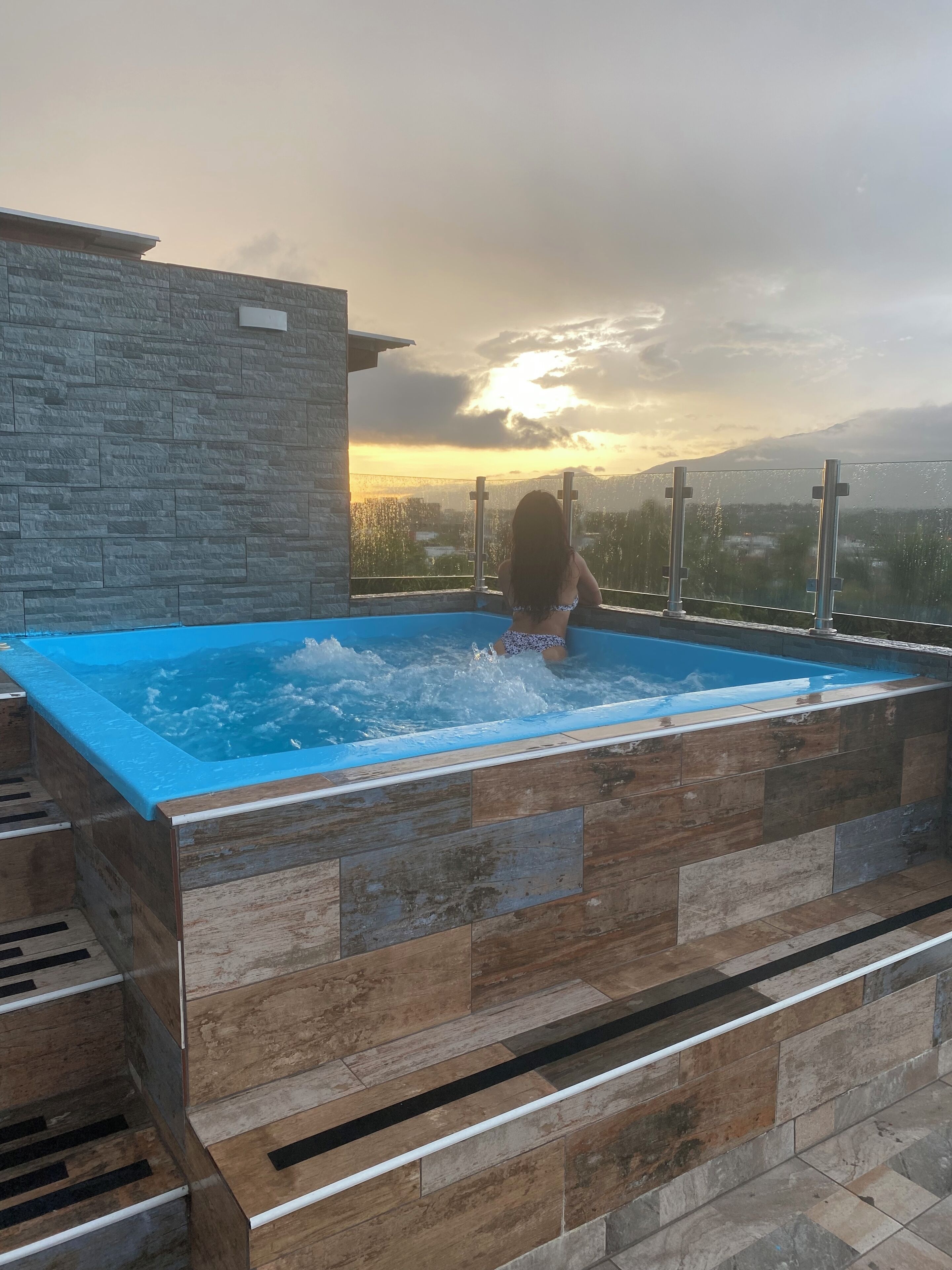 A person in a swimming pool with a view of a sunset and mountains in the background. - Apartment with jacuzzi (Santiago De Los Caballeros)