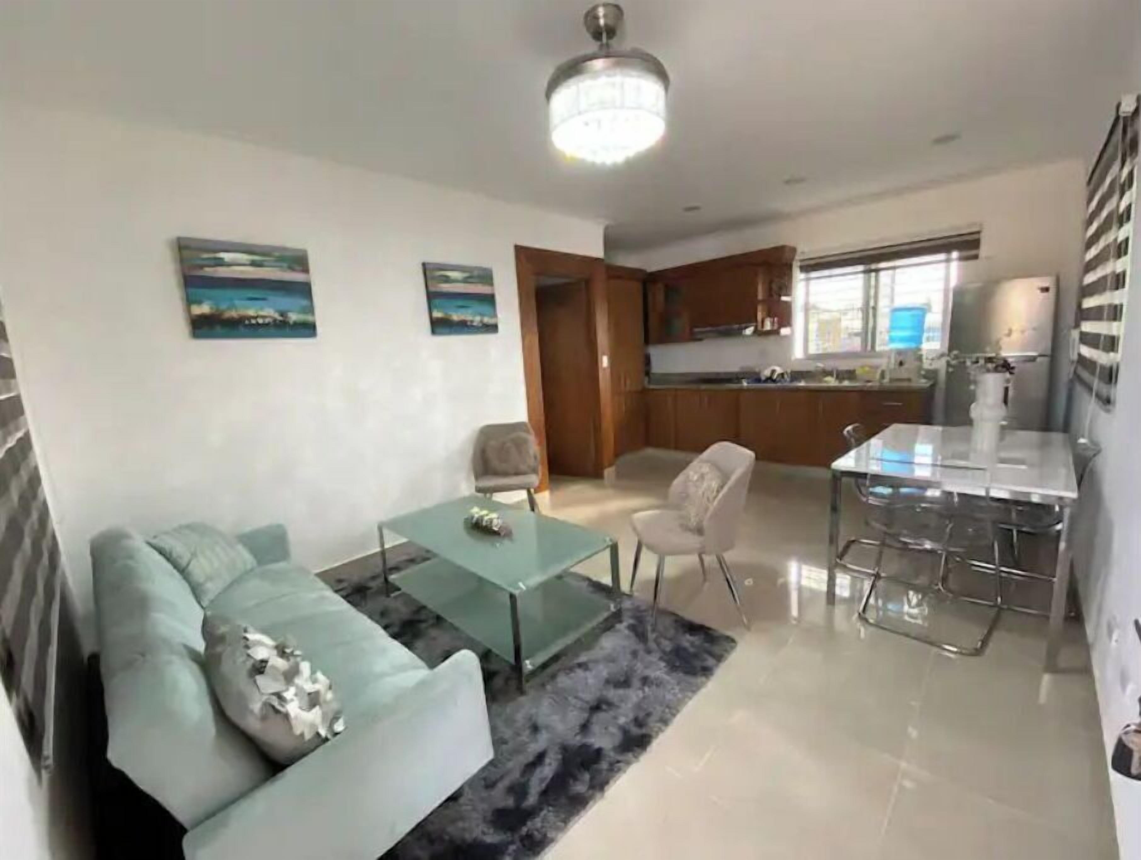 A modern living room with a glass coffee table, a sofa, and a dining area with a glass table and chairs. There is a kitchen in the background with wooden cabinets and a window.