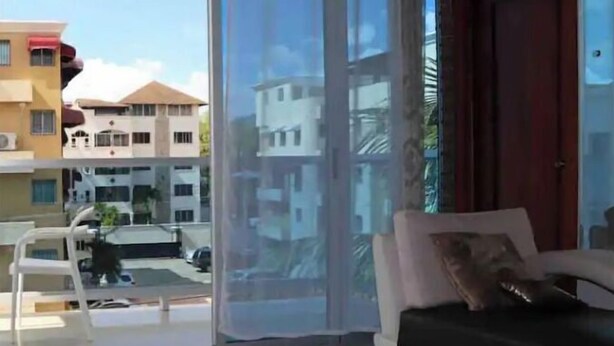 Een moderne woonkamer met een glazen deur naar een balkon, een witte bank en uitzicht op omliggende gebouwen.