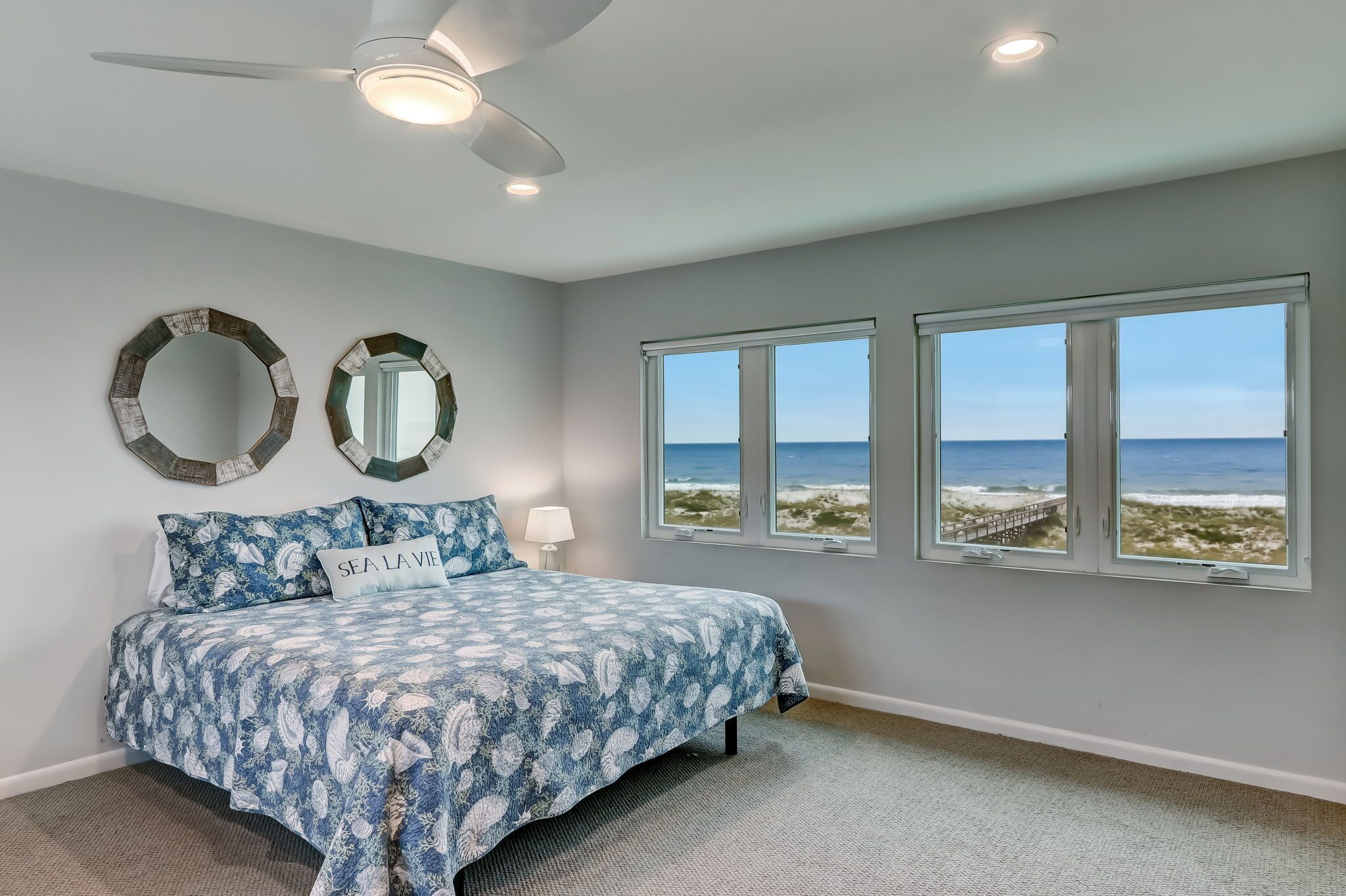 Un dormitorio con cama, dos espejos en la pared, ventilador de techo y vista a la playa a través de amplias ventanas.