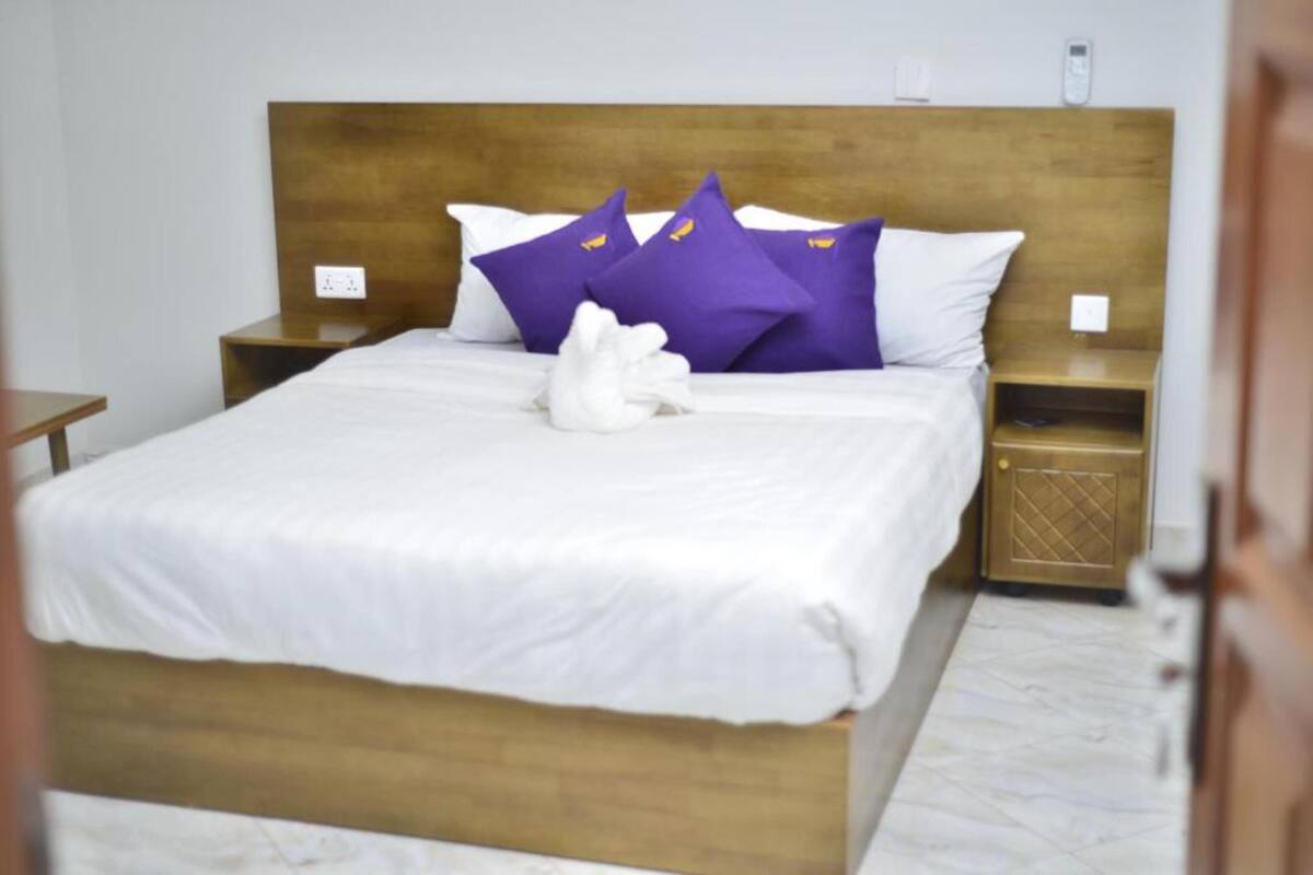 A neatly made bed with white bedding and purple pillows, a wooden headboard, and a small bedside table with a lamp.