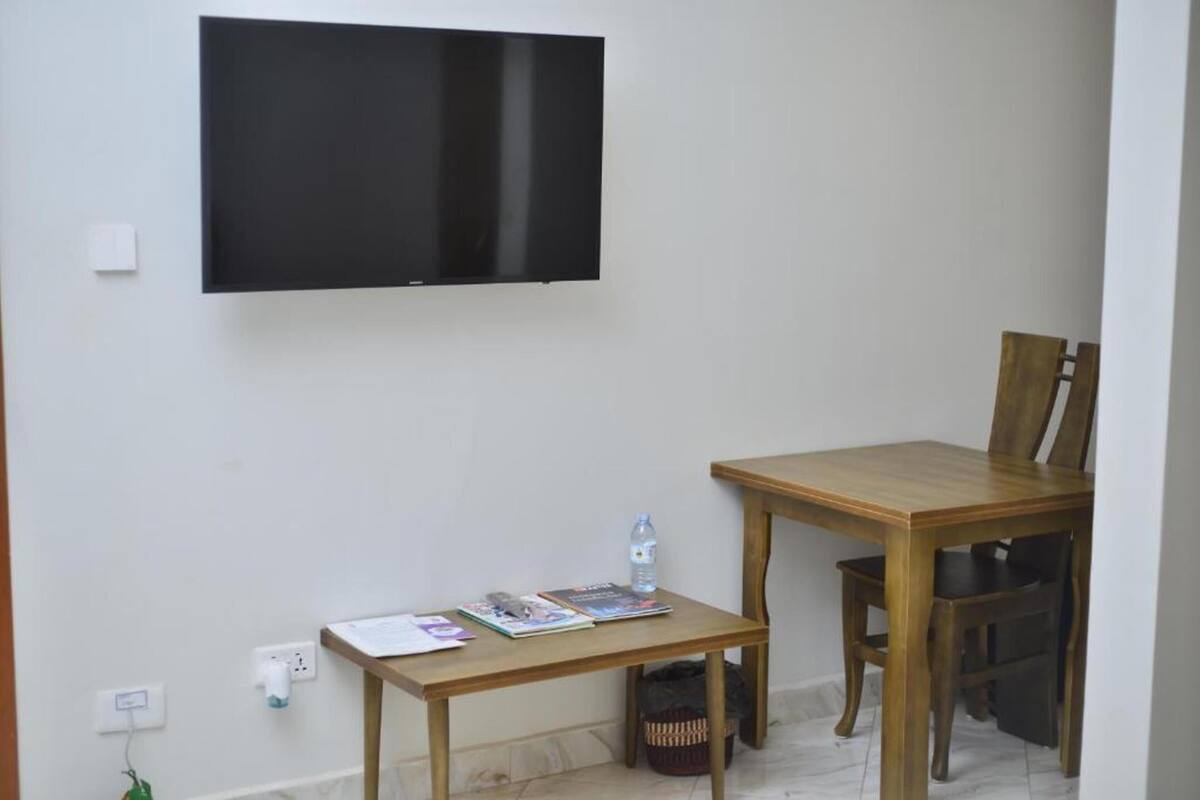 A room with a flat-screen TV mounted on the wall, a wooden table with chairs, and a small side table with a water bottle and magazines.