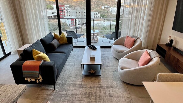 A modern living room with a sofa, armchairs, a coffee table, and a TV. Large windows offer a view of buildings and hills. - New! Slick designer apARTtment with bay views. (Cabo San Lucas)
