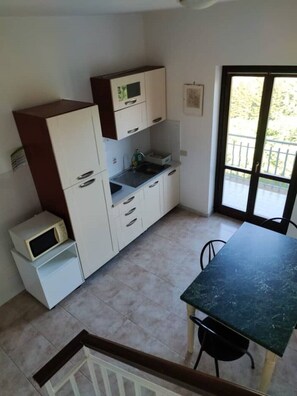 Une cuisine compacte avec des meubles blancs, un four à micro-ondes et un évier. On y trouve une table à manger avec des chaises, et une porte-fenêtre coulissante offre une vue sur l’extérieur.