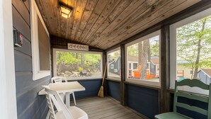 Une véranda couverte avec un plafond en bois, des chaises en plastique blanches, un banc vert et une vue sur un quartier résidentiel avec des maisons et des arbres.