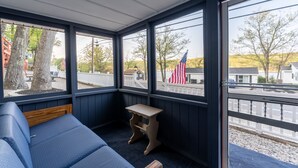 A corner room with a blue sofa, a small wooden table, and large windows offering a view of trees and a white fence. - Delightful Lake View Cabin in Alton Bay Now featuring Lake Access, AC/ TV/ WiFi (Alton)